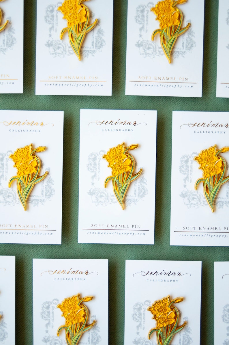 May include: A set of eight yellow daffodil enamel pins on white cards with the text "Seniman Calligraphy" and "Soft Enamel Pin" printed on them. The cards are arranged in a grid pattern on a green surface.
