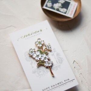 May include: A white enamel pin featuring a delicate sprig of white flowers with green leaves. The pin is on a white card with the text "Soft Enamel Pin" and "JeniMar Calligraphy.com".
