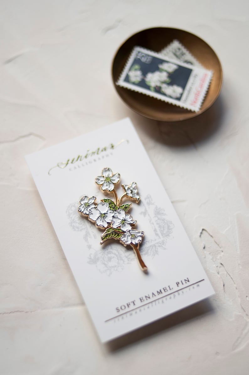 May include: A white enamel pin featuring a delicate sprig of white flowers with green leaves. The pin is on a white card with the text "Soft Enamel Pin" and "JeniMar Calligraphy.com".