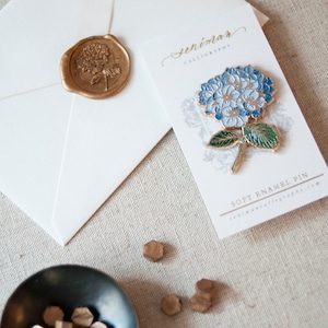 K&ouml;nnte beinhalten: Ein wei&szlig;er Umschlag mit einem goldenen Siegel aus Wachs, das ein Blumenmuster zeigt. Daneben befindet sich eine wei&szlig;e Karte mit dem Text "senimar calligraphy" und einer blauen und gr&uuml;nen Emaille-Anstecknadel mit einer Hortensienbl&uuml;te. Die Karte tr&auml;gt au&szlig;erdem den Text "soft enamel pin" und "senimarcalligraphy.com".