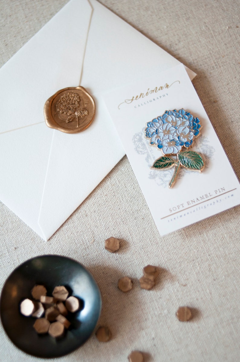 K&ouml;nnte beinhalten: Ein wei&szlig;er Umschlag mit einem goldenen Siegel aus Wachs, das ein Blumenmuster zeigt. Daneben befindet sich eine wei&szlig;e Karte mit dem Text "senimar calligraphy" und einer blauen und gr&uuml;nen Emaille-Anstecknadel mit einer Hortensienbl&uuml;te. Die Karte tr&auml;gt au&szlig;erdem den Text "soft enamel pin" und "senimarcalligraphy.com".