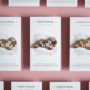 May include: Soft enamel pins featuring a gold-colored outline of a state, adorned with white and green floral accents. Each pin is displayed on a white card with the text "SOFT ENAMEL PIN" and the seller's website.