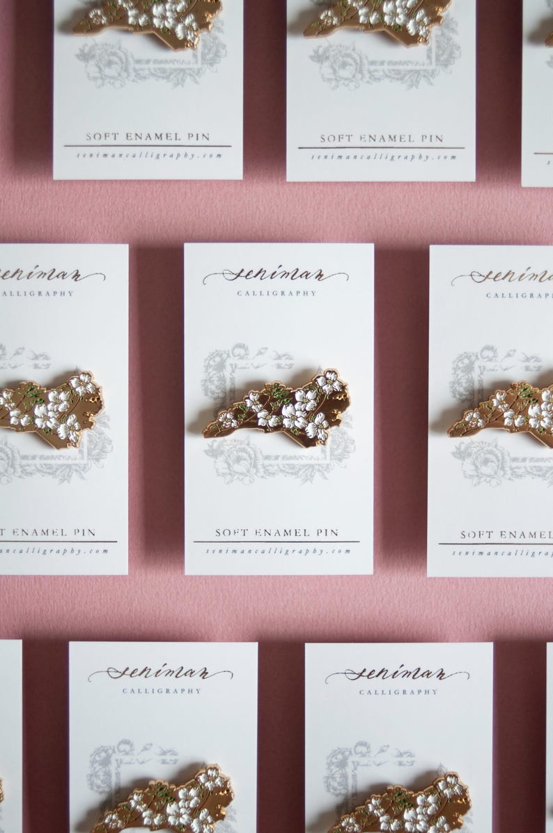 May include: Soft enamel pins featuring a gold-colored outline of a state, adorned with white and green floral accents. Each pin is displayed on a white card with the text "SOFT ENAMEL PIN" and the seller's website.