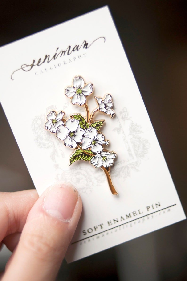 May include: A white enamel pin featuring a delicate floral design with five white flowers and green leaves. The pin is on a white card with the text "Seniman Calligraphy" and "Soft Enamel Pin" in black.