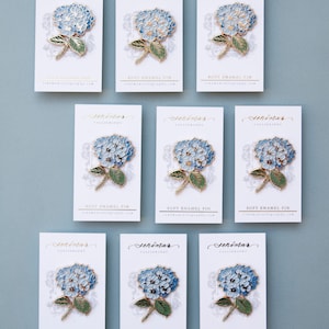 K&ouml;nnte beinhalten: Neun wei&szlig;e Karten mit goldener Beschriftung und blauen und goldenen Hortensien-Blumennadeln. Die Karten zeigen "Senimar Calligraphy" und "Soft Enamel Pin".