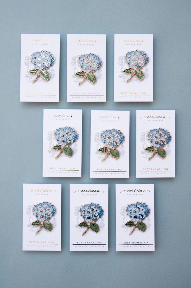 K&ouml;nnte beinhalten: Neun wei&szlig;e Karten mit goldener Beschriftung und blauen und goldenen Hortensien-Blumennadeln. Die Karten zeigen "Senimar Calligraphy" und "Soft Enamel Pin".