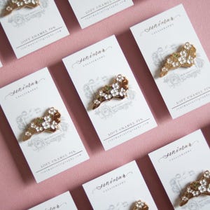 May include: Soft enamel pins with a floral design are displayed on white cards against a pink background. The pins have a gold-colored base with white and green accents. The cards feature calligraphy and the text "SOFT ENAMEL PIN".
