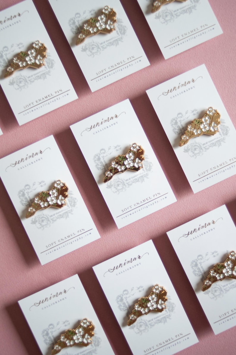 May include: Soft enamel pins with a floral design are displayed on white cards against a pink background. The pins have a gold-colored base with white and green accents. The cards feature calligraphy and the text "SOFT ENAMEL PIN".