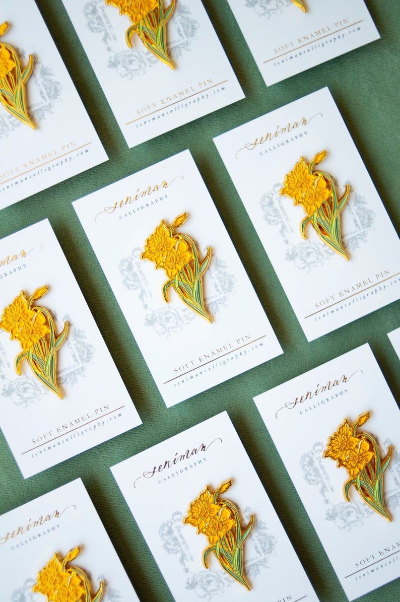 May include: A set of eight white cards with gold lettering and a yellow daffodil enamel pin on each card. The cards have the text "Seniman Calligraphy" and "Soft Enamel Pin" printed on them.