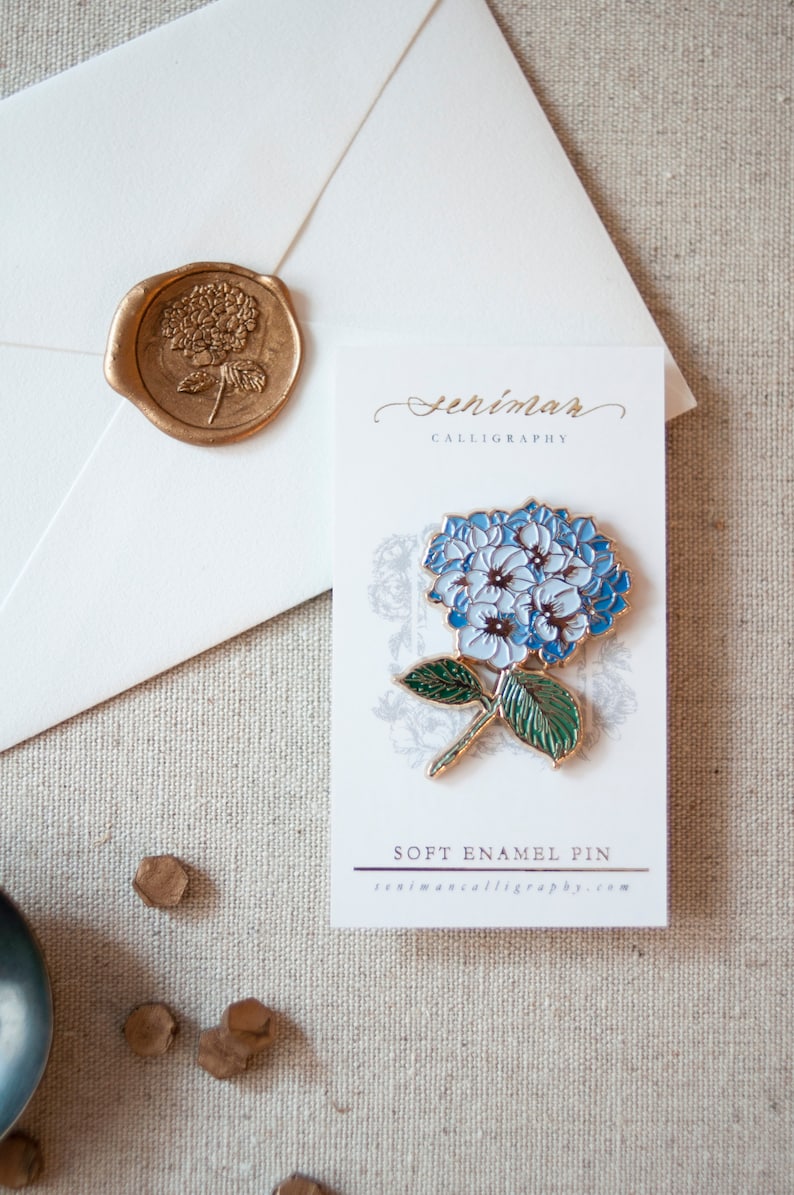 K&ouml;nnte beinhalten: Eine blaue und wei&szlig;e Emaille-Anstecknadel mit einer Hortensienbl&uuml;te und gr&uuml;nen Bl&auml;ttern. Die Anstecknadel befindet sich auf einer wei&szlig;en Karte mit dem aufgedruckten Text "Senimar Calligraphy" und "Soft Enamel Pin".
