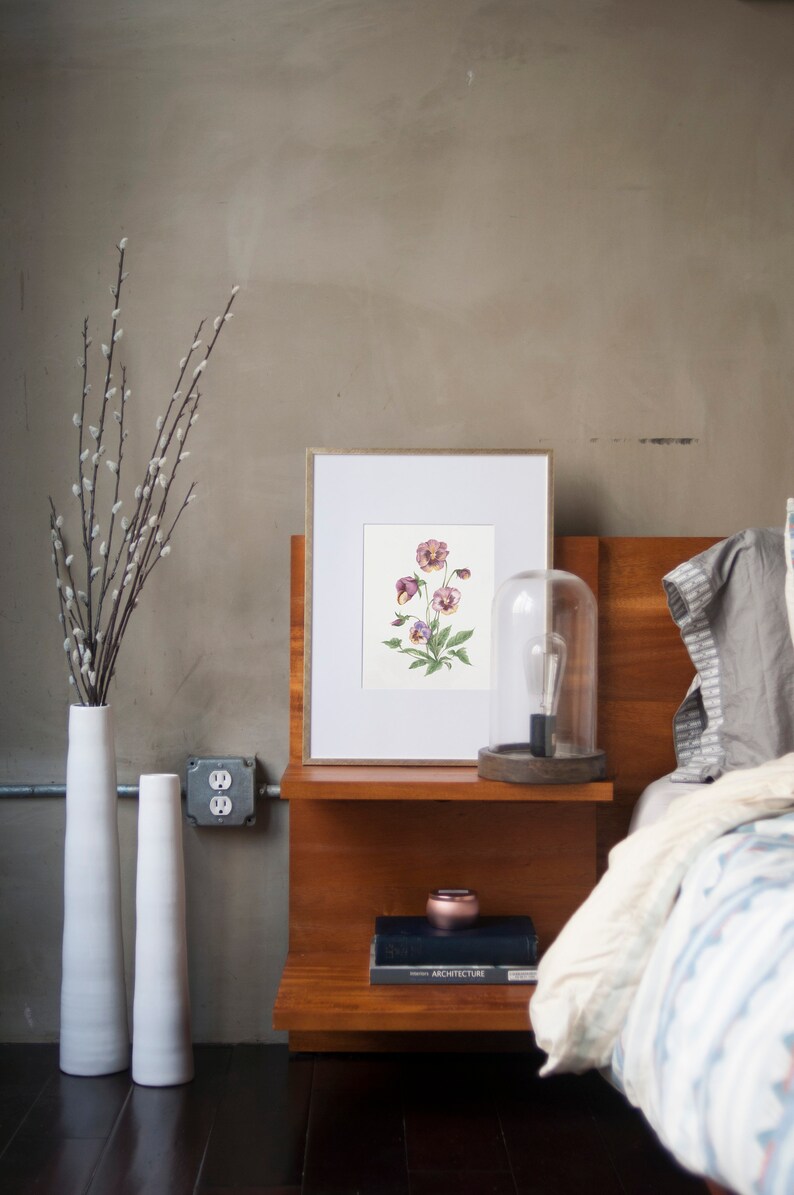 May include: A framed botanical print of purple and white pansies with green leaves on a white background. The print is on a wooden nightstand with a book and a small decorative object. There is a clear glass cloche with a light bulb on the nightstand.