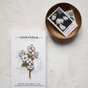 May include: A white and gold enamel pin featuring a delicate white flower with green leaves. The pin is displayed on a white card with the text "Seniman Calligraphy" and "Soft Enamel Pin" printed on it. The card is sitting on a white textured surface.