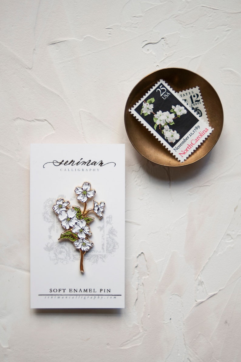 May include: A white and gold enamel pin featuring a delicate white flower with green leaves. The pin is displayed on a white card with the text "Seniman Calligraphy" and "Soft Enamel Pin" printed on it. The card is sitting on a white textured surface.