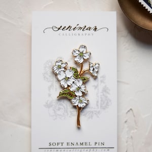 May include: A white enamel pin featuring a gold-toned branch with white flowers and green leaves. The pin is on a white card with the text "Seniman Calligraphy" and "Soft Enamel Pin" printed on it.