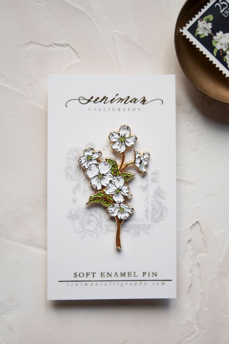 May include: A white enamel pin featuring a gold-toned branch with white flowers and green leaves. The pin is on a white card with the text "Seniman Calligraphy" and "Soft Enamel Pin" printed on it.