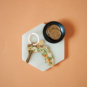 K&ouml;nnte beinhalten: Ein wei&szlig;es, sechseckiges Tablett aus Marmor mit einem goldenen Schl&uuml;sselanh&auml;nger, der die Form des Bundesstaates Florida mit Orangenbl&uuml;ten und einem kleinen goldenen Anh&auml;nger zeigt. Eine schwarze Schale mit goldenen M&uuml;nzen befindet sich auf dem Tablett.