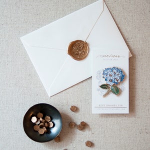 K&ouml;nnte beinhalten: Ein wei&szlig;er Umschlag mit einem goldenen Siegel aus Wachs und einer blauen und gr&uuml;nen Emaille-Blumennadel auf einer Karte mit der Aufschrift "Serendimar Calligraphy" und "Soft Enamel Pin".