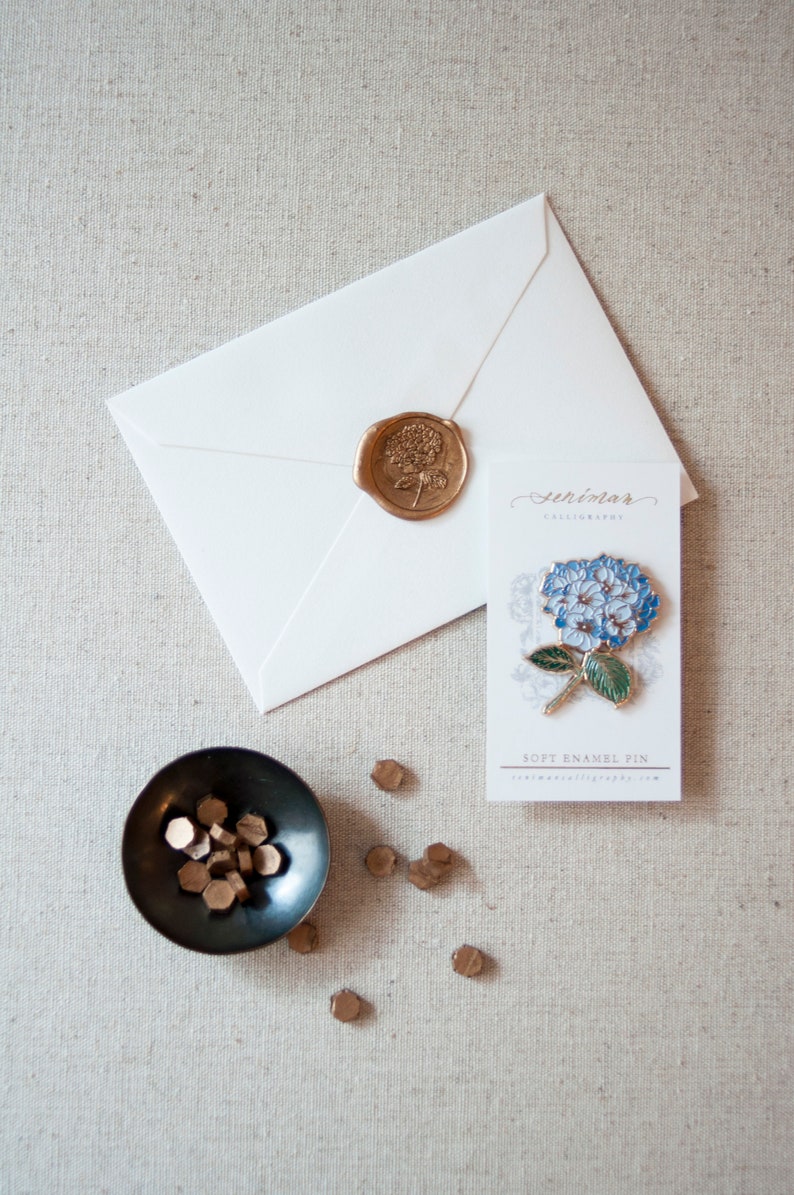 K&ouml;nnte beinhalten: Ein wei&szlig;er Umschlag mit einem goldenen Siegel aus Wachs und einer blauen und gr&uuml;nen Emaille-Blumennadel auf einer Karte mit der Aufschrift "Serendimar Calligraphy" und "Soft Enamel Pin".