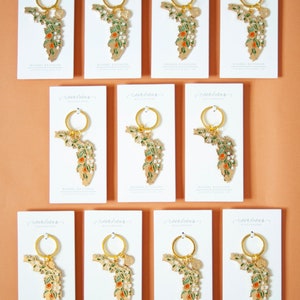 K&ouml;nnte beinhalten: Zw&ouml;lf wei&szlig;e Karten mit goldener Beschriftung und einem goldenen Emaille-Schl&uuml;sselanh&auml;nger mit einem floralen Design des Bundesstaates Florida. Der Schl&uuml;sselanh&auml;nger ist an einem goldenen Ring befestigt. Die Karten tragen die Aufschrift "St. Tropez Calligraphy" und "Enamel Keychain".