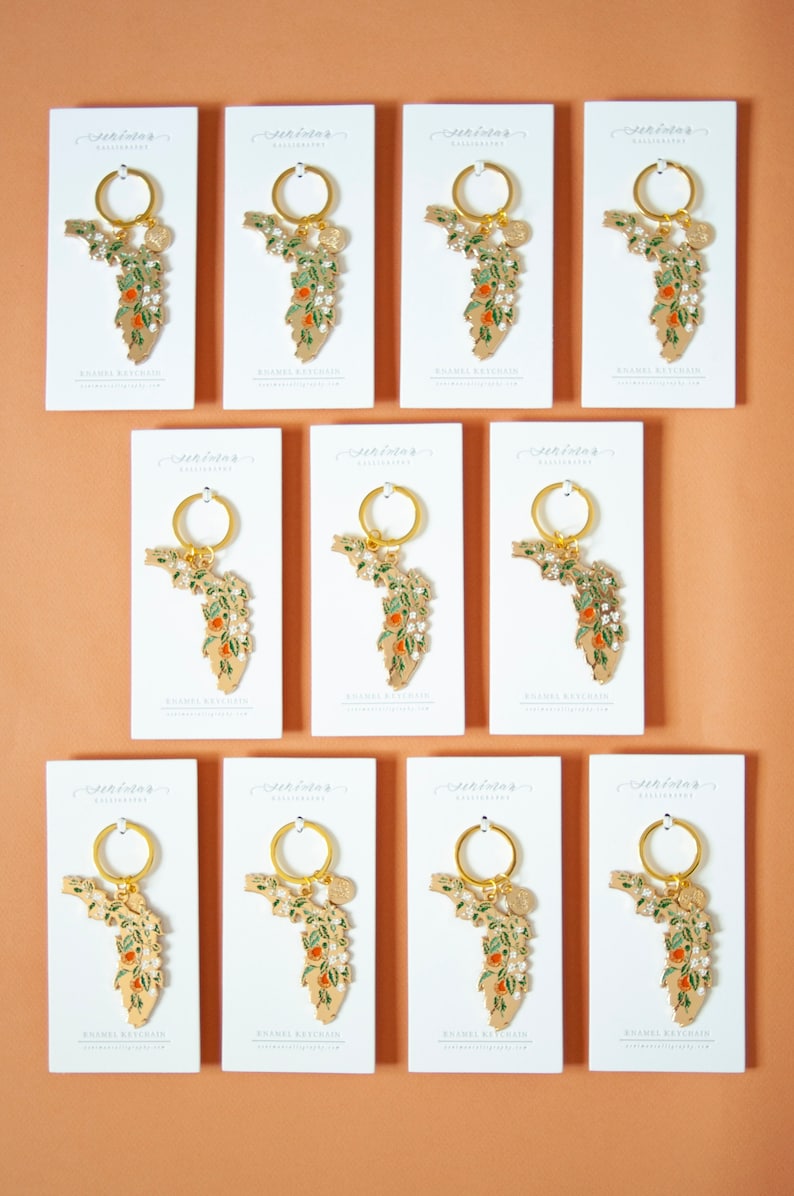 K&ouml;nnte beinhalten: Zw&ouml;lf wei&szlig;e Karten mit goldener Beschriftung und einem goldenen Emaille-Schl&uuml;sselanh&auml;nger mit einem floralen Design des Bundesstaates Florida. Der Schl&uuml;sselanh&auml;nger ist an einem goldenen Ring befestigt. Die Karten tragen die Aufschrift "St. Tropez Calligraphy" und "Enamel Keychain".