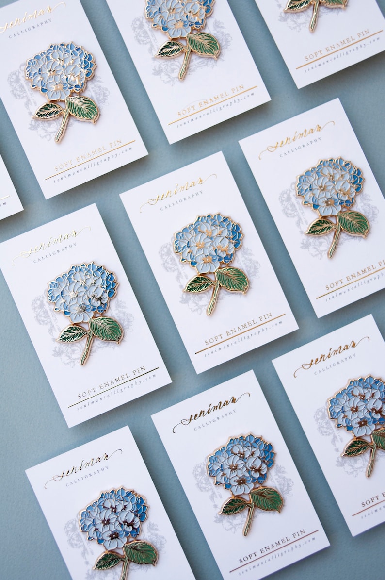 K&ouml;nnte beinhalten: Ein Satz von acht wei&szlig;en Karten mit goldenen Schriftz&uuml;gen und einer blauen und wei&szlig;en Hortensien-Emaille-Anstecknadel auf jeder Karte. Die Karten tragen den Text "Seniman Calligraphy" und "Soft Enamel Pin". Die Anstecknadeln haben alle das gleiche Design und sind in einem Raster angeordnet.