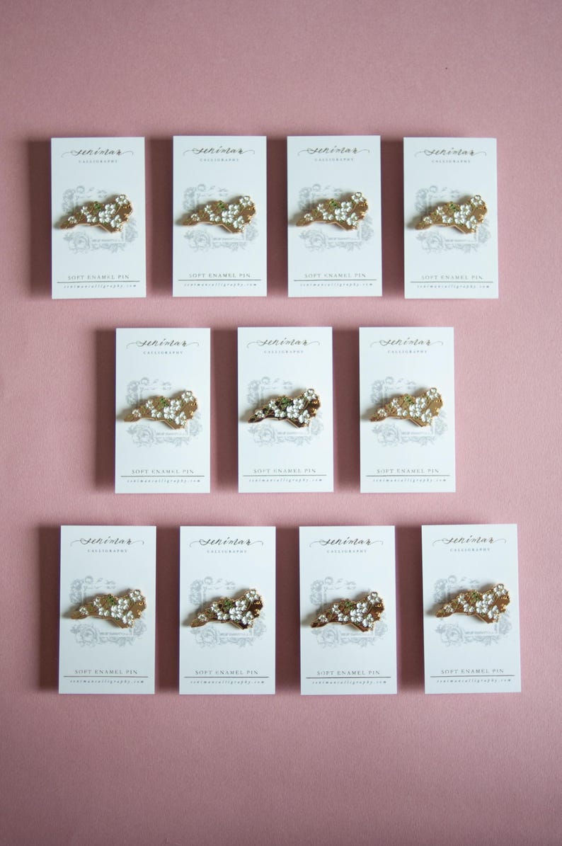 May include: Arrangement of soft enamel pins on white cards against a pink background. Each pin features a gold-colored design with white floral accents. The cards display the text "Sekindar Calligraphy" and "Soft Enamel Pin".