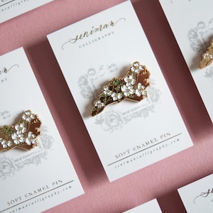 May include: Soft enamel pin featuring a gold-colored outline of a state, adorned with white flowers and green leaves. The pin is displayed on a white card with the text "Seniman Calligraphy" and "Soft Enamel Pin". The background is a soft pink.