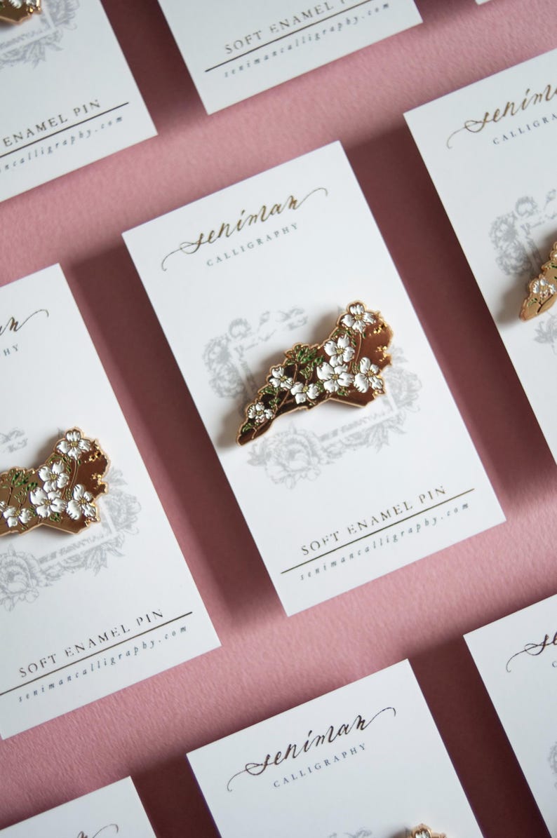 May include: Soft enamel pin featuring a gold-colored outline of a state, adorned with white flowers and green leaves. The pin is displayed on a white card with the text "Seniman Calligraphy" and "Soft Enamel Pin". The background is a soft pink.