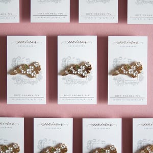 May include: Rows of soft enamel pins on white cards with the word "Seniman" and "Calligraphy" printed at the top. The pins are gold-colored with white and green accents. The cards are on a pink background.