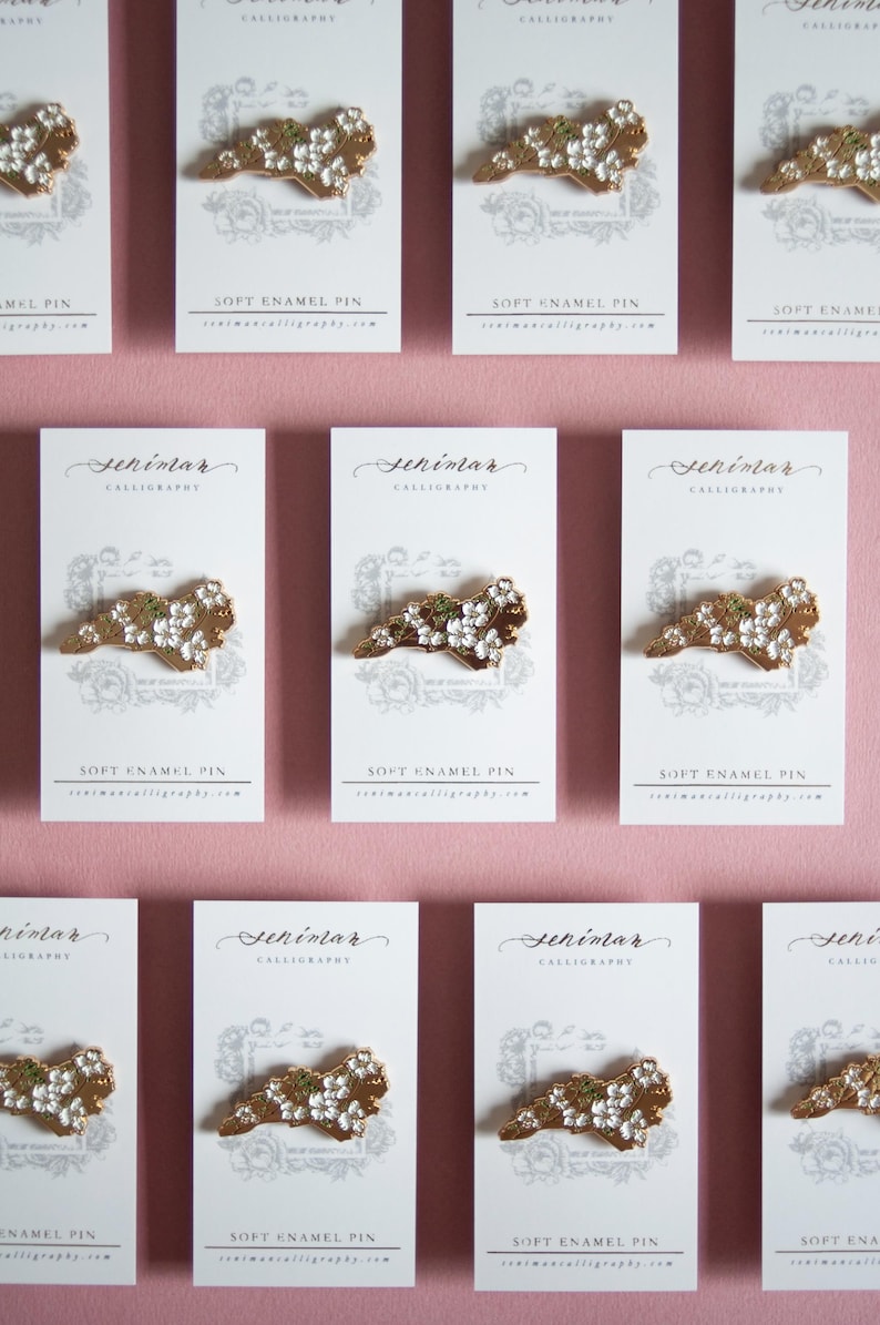 May include: Rows of soft enamel pins on white cards with the word "Seniman" and "Calligraphy" printed at the top. The pins are gold-colored with white and green accents. The cards are on a pink background.