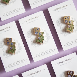 Può includere: Una serie di spille in smalto morbido, ognuna con un contorno dorato di uno stato, adornata con fiori blu e foglie verdi. Le spille sono esposte su biglietti bianchi con calligrafia e la scritta "SOFT ENAMEL PIN".