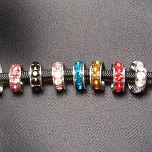 May include: Ten different colored metal rings with clear crystal embellishments. The rings are strung on a black cord.