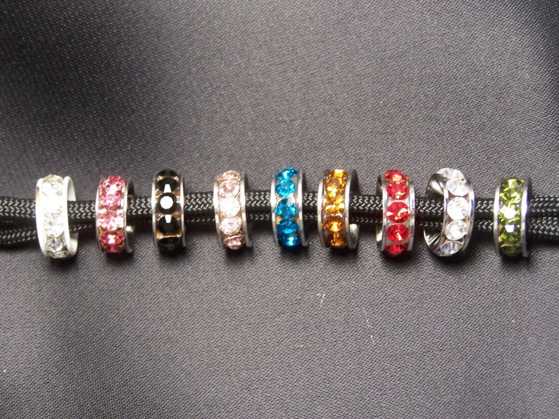 May include: Ten different colored metal rings with clear crystal embellishments. The rings are strung on a black cord.