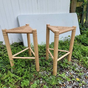 May include: Two wooden stools with triangular seats woven with natural fiber rope. The stools are standing on a gravel surface with green grass and plants.