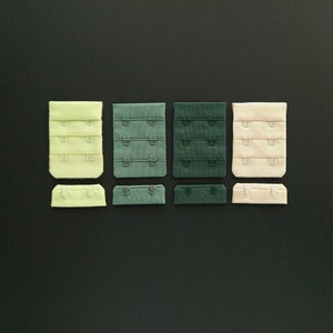 May include: Three sets of bra hook and eye closures in different colours: light green, dark green, and beige. Each set has three rows of hooks and eyes.