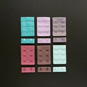 Könnte beinhalten: Neun verschiedene BH-Trägerverlängerungen in verschiedenen Farben, darunter Türkis, Lila, Braun und Rosa. Die Verlängerungen sind in einem Rastermuster auf einem schwarzen Hintergrund angeordnet.