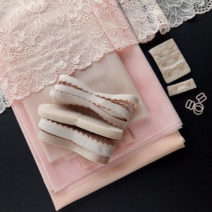 May include: A set of pink fabric pieces for sewing a bra, including lace, mesh, and elastic. The fabric pieces are laid out on a black surface, along with bra clasps and hardware.