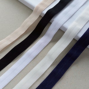 May include: Close-up of several elastic bands in various colors. The bands include beige, black, white, off-white, and navy blue. The bands are arranged diagonally across the frame, showcasing their texture and color variations. These bands are likely used for sewing or crafting projects.