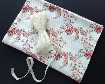 May include: A white fabric with a floral pattern and thin brown stripes. A white spool of ribbon and a white lace ribbon are on top of the fabric.