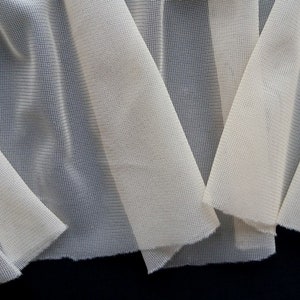 Puede incluir: Primer plano de un trozo de tela transparente de color blanco roto con textura tejida. La tela est&aacute; doblada en varias capas, creando un efecto de drapeado suave.