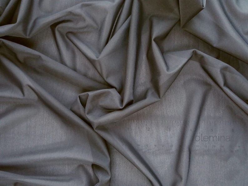 May include: A close-up of a smooth, dark gray fabric with a subtle texture. The fabric is draped and folded, creating soft, flowing lines.