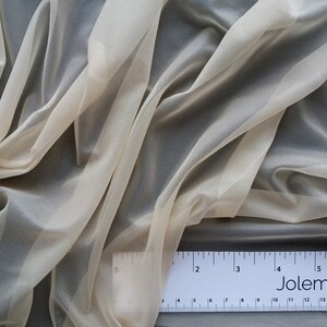 Puede incluir: Un primer plano de una tela transparente de color marfil con un brillo sutil. La tela est&aacute; drapeada y doblada, revelando su textura delicada. Una regla con la palabra "Jolemina" impresa en ella est&aacute; colocada sobre la tela.