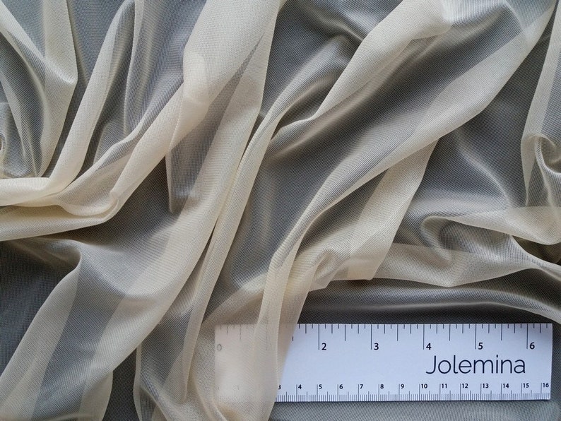 Puede incluir: Un primer plano de una tela transparente de color marfil con un brillo sutil. La tela est&aacute; drapeada y doblada, revelando su textura delicada. Una regla con la palabra "Jolemina" impresa en ella est&aacute; colocada sobre la tela.
