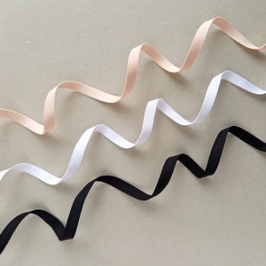May include: Three different colored ribbons, black, white, and light pink, are arranged in a wavy pattern on a light gray background.