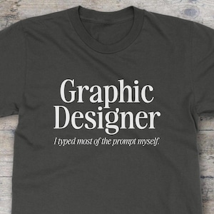 以下が含まれることがあります： チャコールグレーのクルーネックTシャツ。中央には大きなセリフ体で「Graphic Designer」と白文字でプリントされ、そのすぐ下には「I typed most of the prompt myself」という小さな白い文字が、すっきりとしたイタリック体のサンセリフフォントで配置されています。