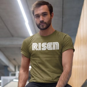 Op de afbeelding: Olijfgroen t-shirt met het woord "RISEN" in witte blokletters. Het shirt heeft korte mouwen en een ronde hals. De tekst staat in het midden op de borst. Het model draagt een zwarte broek.