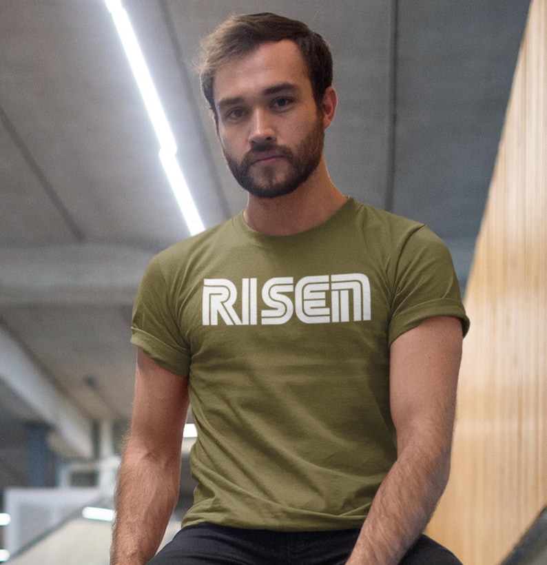 Op de afbeelding: Olijfgroen t-shirt met het woord "RISEN" in witte blokletters. Het shirt heeft korte mouwen en een ronde hals. De tekst staat in het midden op de borst. Het model draagt een zwarte broek.