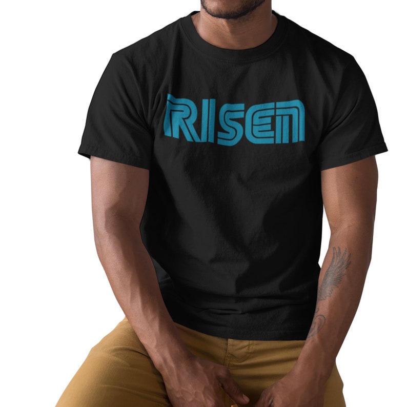 Op de afbeelding: Zwart T-shirt met het woord "RISEN" in een turquoise, retro-stijl lettertype over de borst. Het shirt heeft korte mouwen en lijkt van een zacht materiaal te zijn. De persoon die het shirt draagt, zit.