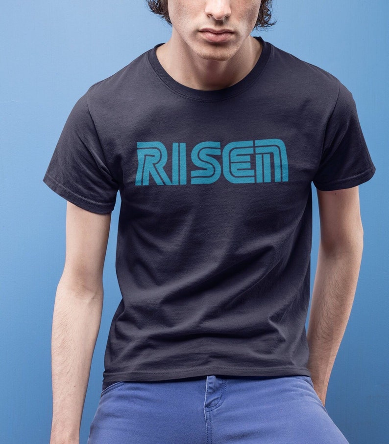 Op de afbeelding: Een marineblauw t-shirt met het woord "RISE" in turquoise blokletters. De tekst heeft een retro, gelaagd ontwerp. Het shirt met korte mouwen lijkt van een zacht materiaal te zijn.