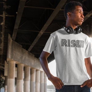 Op de afbeelding: Een wit t-shirt met het woord "RISEN" in grijs gedrukt, met een retro lettertype. Het shirt met korte mouwen wordt gedragen door een persoon met een koptelefoon om de nek. De achtergrond bevat betonnen pilaren en balken.